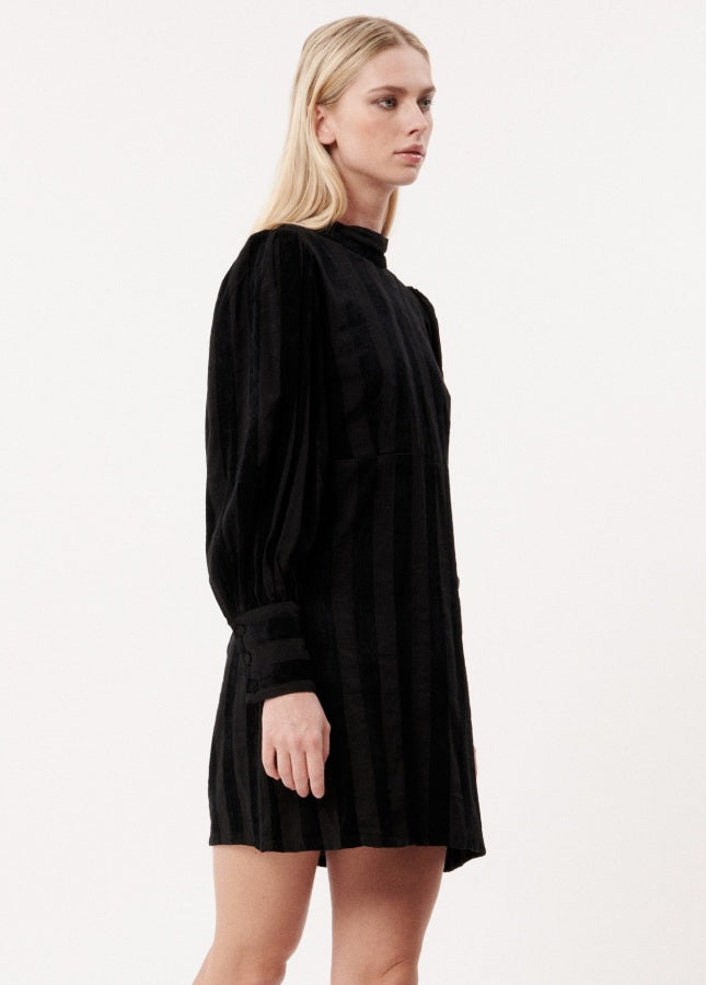 Zwarte midi-jurk met open rug Cecilia van het merk FRNCH. De zwarte jurk heeft subtiele verticale fluwelen strepen. De midi-jurk heeft lange (lichte) pofmouwen met manchetten aan de uiteinde en een diepe V-hals op de rug met strik in de nek. Shop de nieuwe collectie jurken, zwarte jurken, FRNCH jurken bij Boetiek Aniek