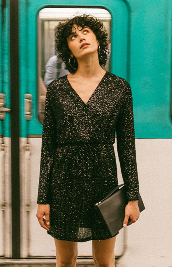 Zwarte paillettenjurk Love van het merk Grace & Mila. De zwarte korte pailletenjurk heeft een V-hals (met drukknoopje), lange mouwen en een elastische tailleband. De paillettenjurk heeft een zwarte onderjurk. De perfecte zwarte jurk voor een feestje of voor tijdens de feestdagen. Shop paillettenjurken online.